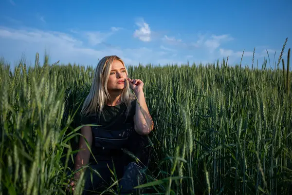 stock image In the tranquil expanse of a rustic wheat field, a female farmer squats, holding a wheat stalk in one hand, contemplatively placing it near her lips. With sun-kissed skin and clad in work attire, she