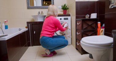 İnce vücutlu, pembe kazak ve mavi kot giyen bir kadın, çamaşır makinesinin yanında, parlak, rahat bir banyoda oturur, yeni yıkanmış bluzunu leke var mı diye dikkatle kontrol eder..