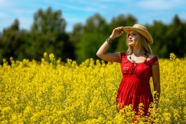 Canlı kırmızı elbiseli bir kadın zarif bir şekilde sarı bir çiçek tarlasında duruyor, güneşli günün tadını çıkarıyor.