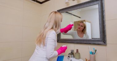 Pembe eldiven takan bir kadın banyo aynasını özenle temizler. Ev hijyeninin önemini göstermekten mutluluk duyar.
