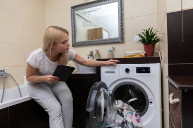 Bir kadın şık bir çamaşır odasında bulunan modern çamaşır makinesinin çeşitli ayarlarını dikkatlice kontrol ediyor.