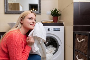Sıcak bir çamaşır odasında, bir kadın neşeyle gülümsüyor, çamaşır makinesinin yanında yeni yıkanmış bir havlu tutuyor.