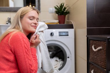 Bir kadın çamaşır makinesinin yanında dururken temiz çamaşırların canlandırıcı ve taze kokusundan zevk alır.