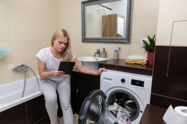 Genç bir kadın şık ve çağdaş bir banyoda çamaşır yıkarken tabletinin keyfini çıkartıyor.