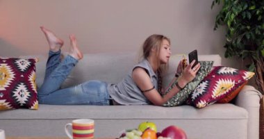 Genç bir kadın rahat bir şekilde kanepede uzanıyor, rahat bir yaşam alanında akıllı telefonuyla meşgul.