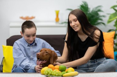 Evde çeşitli meyvelerle çevriliyken çocukların köpekleriyle mutlu mesut oynamalarının iç açıcı ve neşeli bir sahnesi.