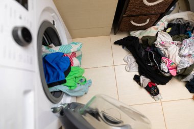 Çamaşır makinesi, yerdeki bir yığın çamaşırın yanına yerleştirilmiş renkli giysilerle doludur.