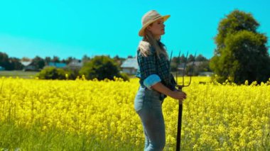 Bir kadın canlı sarı bir tarlada yürür, gururla bir yaba tutar ve kırsal yaşamı sergiler.