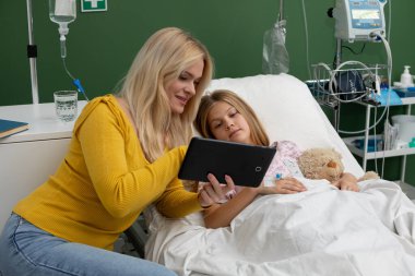 Şefkatli bir anne, hasta kızını renkli bir tabletle eğlendirir ve moralini yüksek tutar.