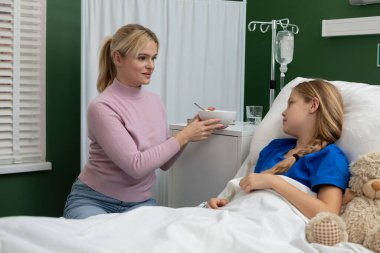 Sıcak bir hastane odasında, kendini adamış bir anne hasta kızını şefkatle besliyor, sevgi ve ilgi gösteriyor.