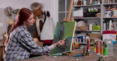 Odaklanmış bir sanatçı yaratıcı stüdyosunda çeşitli sanat malzemeleriyle dolu canlı bir tuval çizer.