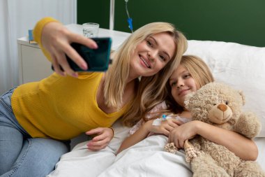 Sevgi dolu bir anne ve kızı, bir hastane odasında keyifli bir selfie çekerken neşe dolu bir anı paylaşıyorlar.