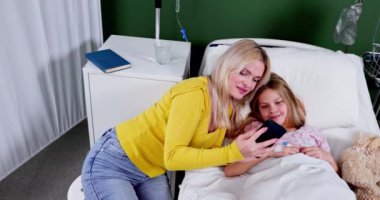 Bir anne ve kızı, hastane odasında rahat etmek için mobil bir cihaz kullanarak samimi bir anı paylaşıyor.
