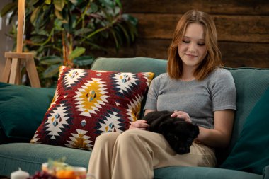 Bir kadın sıcak, davetkar bir oturma odasında siyah bir kedi kıvrılıp yatarken rahat koltuğunda biraz dinlenir.