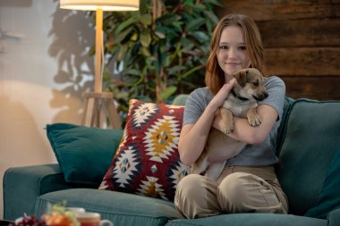 Bir kadın sıcak ve davetkar bir atmosfer yaratmak için rahat bir kanepede küçük bir köpeği kucaklarken zevkle ışıldıyor.