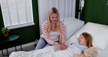 Şefkatli bir anne, hasta kızını sıcak ve derin bir ilgi göstererek, parlak bir hastane odasında sevgiyle rahatlatır.