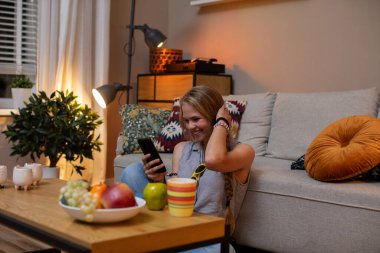 Sıcak ve rahat bir iç mekan sahnesinde bir insan akıllı telefonunu kullanırken rahat bir şekilde meyve yiyor.