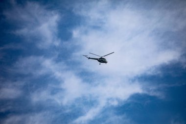 Bir askeri helikopter, tüylü beyaz bulutlarla kaplı açık mavi bir gökyüzünde ustalıkla manevra yapar.