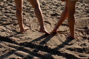 Güneşli bir kumsalda ardında neşeli desenler bırakarak kumda konumlanmış iki çift ayağın detaylı bir yakın çekim görüntüsü.