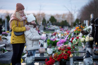 Huzurlu bir mezarlıkta, bir anne ve çocuğu arasında canlı çiçeklerin arasında dokunaklı bir hatıra anı belirir.