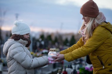 Son derece dokunaklı bir sahne, gün batımında huzurlu bir mezarlıkta parlayan bir fener tutan bir kadın ve çocuğu resmediyor.