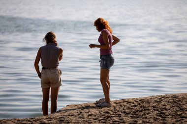 İki kadın dingin suyun yanında huzur içinde durmuş kumsalda güneşli ve güzel bir günün tadını çıkarıyorlar.