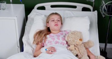 A young girl lies in a hospital bed, holding her teddy bear, symbolizing her vulnerability and innocence