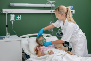 Kendini adamış bir sağlık çalışanı, tedavi için maske takan genç bir kıza hastane yatağında destek olur.