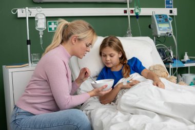 Genç kızını bir hastane yatağında sevgiyle rahatlatan bir annenin yürek ısıtan ve besleyen bir sahnesi.