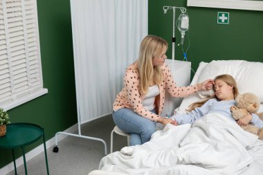 Dokunaklı bir sahne hasta çocuğuna bakan bir anneyi umut dolu sıcak bir hastane odasında gösteriyor.