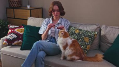 Neşeli bir kadın tüylü turuncu kedisiyle renkli yastıklarla çevrili rahat bir kanepede etkileşime girer.