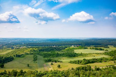 Tüylü beyaz bulutlarla bezenmiş berrak mavi gökyüzünün altında soluk kesici bir yeşillik manzarası.
