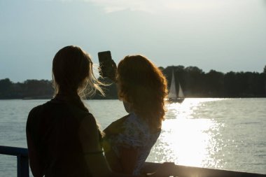 İki arkadaş, arkadaşlıklarını ve maceralarını kutlamak için huzurlu suyun kenarında gün batımında mutlu bir selfie çekiyorlar.