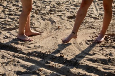 İki çift çıplak ayak, kumlu bir plajda, ılık güneş ışığının altında, yumuşak gölgeler bırakıyor.