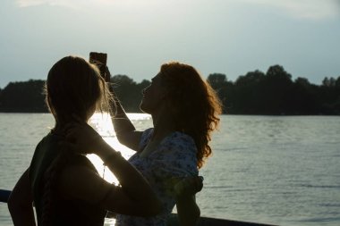Bu huzurlu sahnede, iki arkadaş güzel bir gün batımında parıldayan suyun kenarında neşeyle selfie çekiyorlar.