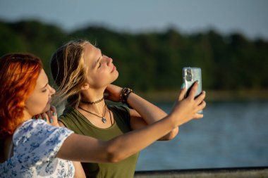 Işıldayan suyun kenarında mutlu bir selfie çeken iki genç kadın, parlak güneşin altında neşeli ve eğlenceli bir anı yakalıyorlar.