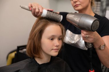 Profesyonel bir stilist kuaförde saç kurutma makinesi ve fırça kullanarak genç bir kızın saçını ustalıkla tarar.
