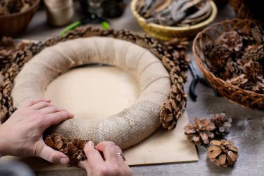 Eller ustalıkla çam kozalakları gibi çeşitli doğal malzemeler kullanarak çarpıcı, güzel ve süslü bir çelenk yaratıyor.