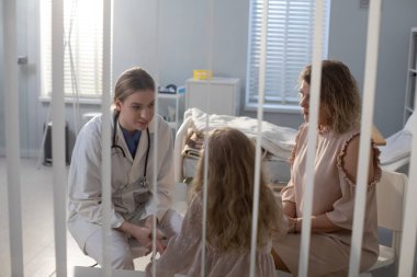 İlgili bir tıbbi uzman, genç bir hasta ve annesiyle hastanede dikkatli bir şekilde iletişim kurar.