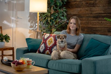 Bir kadın, sıcak bir oturma odasında sevgili köpeğiyle yumuşak bir deniz mavisi kanepede rahatça dinlenirken görülüyor.