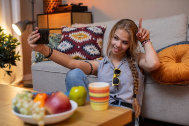 Genç bir kız mutlu bir şekilde evinde parlak bir selfie çekiyor, ön planda samimi bir titreşim ve renkli bir meyve ile çevrili.