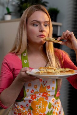 Mutlu bir kadın, neşeli çiçekli bir önlük giyerken canlı mutfağında lezzetli bir dilim pizzanın tadını çıkarıyor.