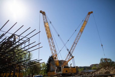 Vinçler yoğun bir inşaat alanında ağır çelik kirişleri kaldırıyor, güneşli havada endüstriyel aktiviteler sergiliyorlar.