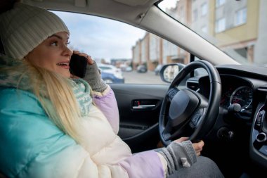 Sıcak kış kıyafeti giymiş bir kadın araba kullanırken telefonla konuşuyor, günlük yaşam tarzını gösteriyor.