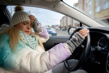 Bir kadın kış kıyafeti giyiyor ve soğuk havada arabasıyla giderken stresli görünüyor.