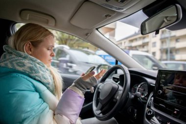 Kışlık montlu genç bir kadın, yavaş giden trafikte beklerken akıllı telefonuna odaklanmış bir şekilde arabada oturuyor.