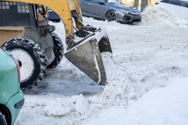 Kar temizleyen bir kar temizleme aracı, kar yığınları ve park halindeki araçlarla çevrili kışın manzarada