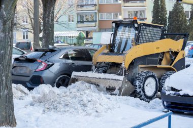 Meşgul bir şehir otoparkında, bir patinaj dümeni yükleyicisi etkin bir şekilde kar temizlerken çok sayıda araç park halinde duruyor.