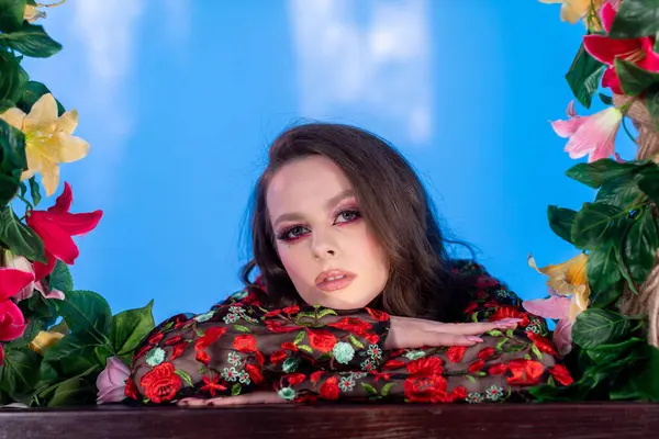 Büyüleyici bir model, sayısız canlı çiçeğin arasında zarif bir şekilde poz veriyor. Serin mavi bir arkaplana kurulmuş.