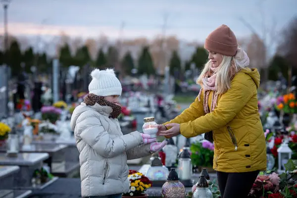 Bir anne ve kızı, huzur dolu bir mezarlıkta, sevdiklerini onurlandıran bir mum yakıyor.